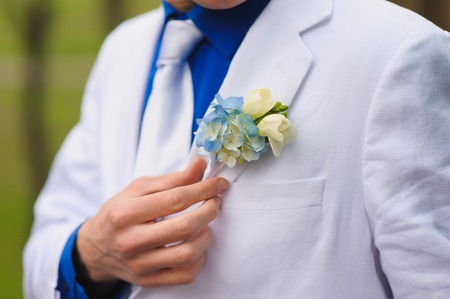 A man in a blue shirt, a white suit and a white tie. Hand corrects boutonniere.の写真素材