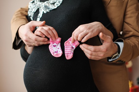 Beautiful pregnant woman and her husband in black dress and brown suit. Husband holds wife's hands with two booties in pregnant belly.の写真素材