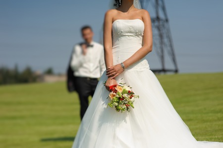 Bride in beautiful dress holding a bouquet of flowers. Blurred groom in the background.の写真素材