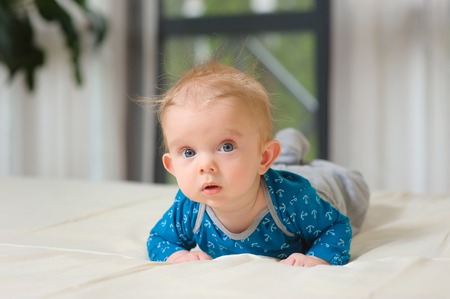 Baby boy lying on belly. Cute year-old kid with interest looks at the camera, boy lying on bed in...