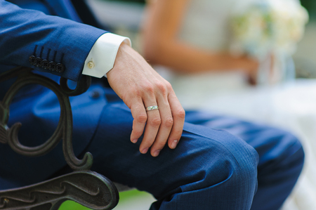 Man, groom in a blue suit sitting on a bench. Close-up. A hand with a ring. Bride with flowers in the background.の写真素材