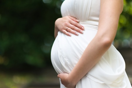 Pregnant young woman. Pregnant woman in white dress relaxing outdoors. Hands on belly, blurred green...