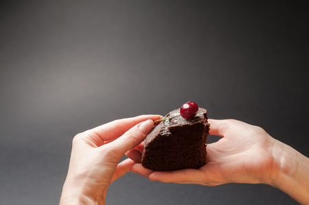 Slice of delicious chocolate cake with cherry on top in a woman hands on a dark background with copy space.の写真素材