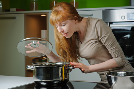 Young woman in the kitchen. Beautiful young housewife looking in to pan. Red-haired girl smiles while preparing food at home kitchen.の写真素材