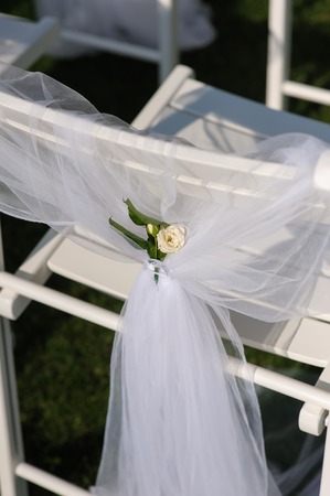 White chair with a small flower. White decorated chairs on a green lawn. Chairs set in rows for the wedding ceremony. They are decorated for the festive event. Chairs are on the green lawn outside.の写真素材