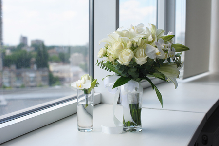 Beautiful wedding bouquet in a glass vase. Flowers on white windowsill. Boutonniere in a small vase. The window in the background.の写真素材