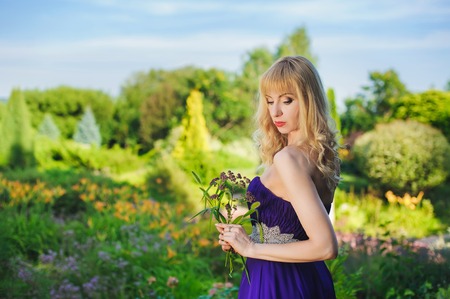 Portrait of beautiful woman dressed in evening gown. Blonde Beautiful woman in long purple dress like princess in green garden. Woman holding bouquet of wildflowers.の写真素材