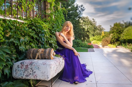 Portrait of beautiful woman dressed in evening gown. Blonde gorgeous woman in long purple luxurious dress posing in garden. Woman sitting on sofa outdoors.の写真素材