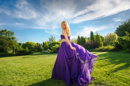 Blonde gorgeous woman in long purple luxurious dress posing in garden. Dress fluttering on wind.の写真素材