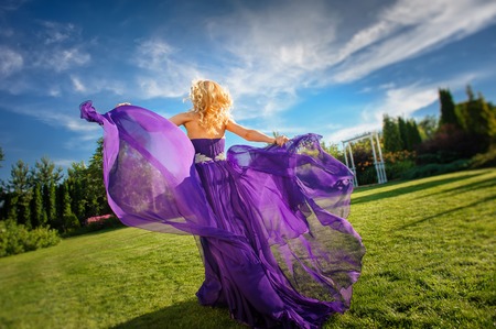 Blonde gorgeous woman in long purple luxurious dress posing in garden. Dress fluttering on wind.の写真素材