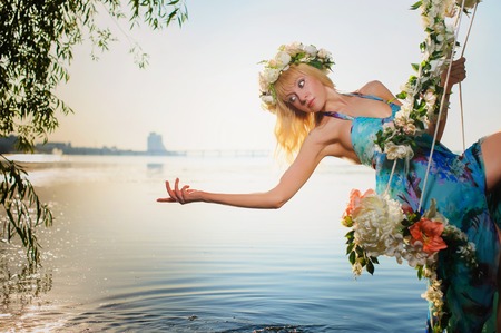 Girl on home made tree swing over river. Swing decorated with flowers. Woman with flower wreath on head.の写真素材
