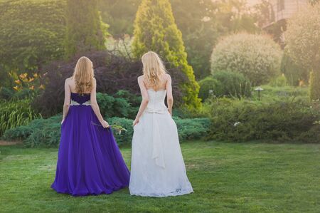 Bride and bridesmaid outside. Two girls posing on the green meadow. Two girls standing back to camera. Bride in wedding dress. Bridesmaid in a purple evening gown.の写真素材