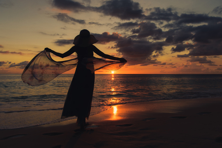 Back view of lonely girl with summer dress and hat is walking along coastline. Silhouette young woman on the beach at sunset. Fluttering dress.の写真素材