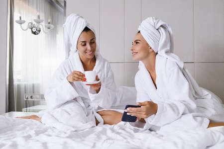 Two females sit at luxury appartment hotel bed at morning with cup, wear bathrobes and towels, look at smartphone, online networking, shopping, chatting, ladies friendship and leisureの写真素材