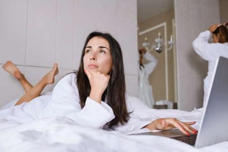 Two girls in beautiful appartment hotel, wear bathrobes after morning shower, check information online using laptop, plan day, business, leisure, vacation togetherの写真素材