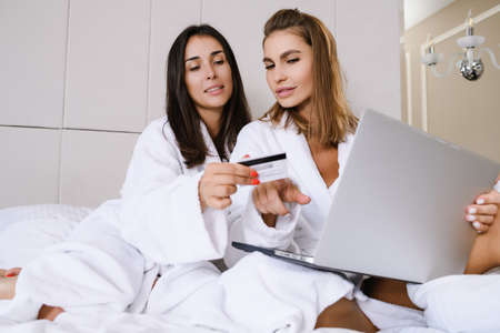 Two young beautiful females in bed wearing white bathrobes hold laptop and bank credit payment card at luxury home, hotel background to order food, services, shopping onlineの写真素材