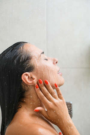 Brunette girl with red manicure polish take shower, bath with tiles background, wash face, hair and neck with gel and water close up.の写真素材