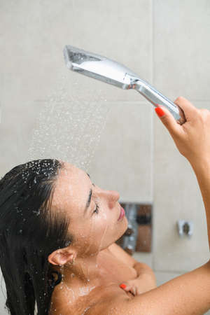 Young woman take shower bath, wash dark hair with faucet up and water drops on beautiful face and...