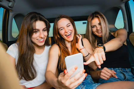 Beautiful female friends have fun, smiling with mobiel phone at vehicle seat back, inside auto salonの写真素材
