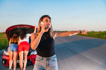 Three confused female friends stuck at empty road, calling to mechanic service, try to fix hood details, stop other vehicles with handの写真素材