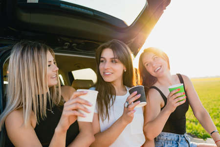 Happy road trip of three smiling female travelers, relax at vehicle back with coffee cups take away at sunlight. The girls are drinking coffee.の写真素材