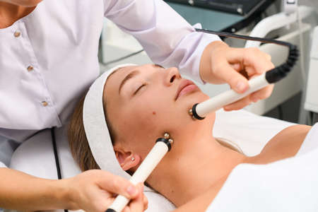 Woman make facial microcurrent myostimulation procedure with rollers at clinic cabinet for client...