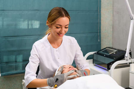 Cosmetologist smile during massage female client face, wearing white robe and gray microcurrent gloves for hardware therapy at beauty clinic cabinetの写真素材