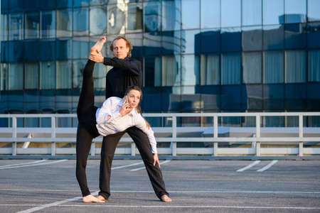 Man and woman make acroyoga element with female leg up, she talk by phone, business and sport concept at modern glass building backgroundの写真素材