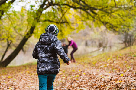 Preteen boy in jacket and hat run to mother and brother at autumn forest background. Active family holiday conceptの写真素材