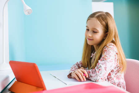 Child primary school age sit at desk with blank exercise book, hold pen and look at tablet screen, study at home during lockdownの写真素材