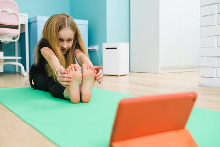 Small girl stretch her legs on mat, looking at tablet screen, online remote sport gymnastics class from home during lockdownの写真素材
