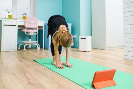 Small girl gymnast in black leotard stay on mat with head down, exercise at home with online lesson using tablet during isolationの写真素材