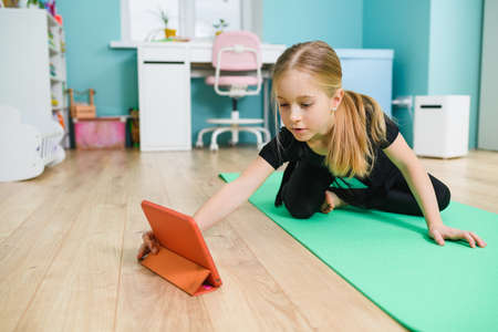 Elementary school girl put sport mat and tablet on floor, wearing black leotard before start online remote gymnastic class during self-isolation at homeの写真素材