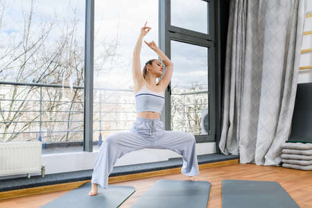 Girl stand in gray sportswear with wide legs on mat and hands up in studio with big window, neutral colors, healthy lifestyleの写真素材