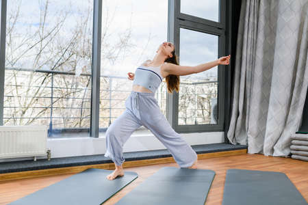 Girl stretching and dancing in light sport studio in gray comfortable sportswear with raising up hand, low angle viewの写真素材