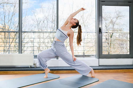 Young woman in cozy gray sportswear stand in freezing posture on mat with wide legs and raised hand up near big windowの写真素材