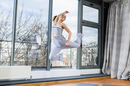 Young woman jumping high barefoot at light studio hall, wearing cozy gray pants and top, healthy active lifestyleの写真素材