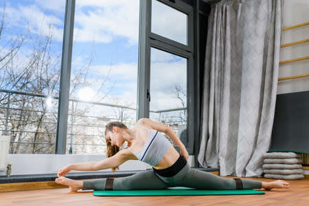Beautiful girl stretching at light studio near panoramic window, sitting in twine and touch foot toes at green mat. Woman bends to the right while sitting in a split.の写真素材