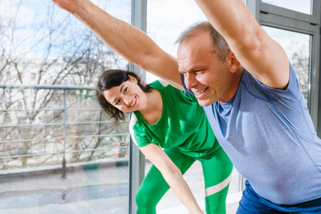 Positive aged man with hands up making wellness training workout with female doctor osteopath indoors, healthy lifestyleの写真素材