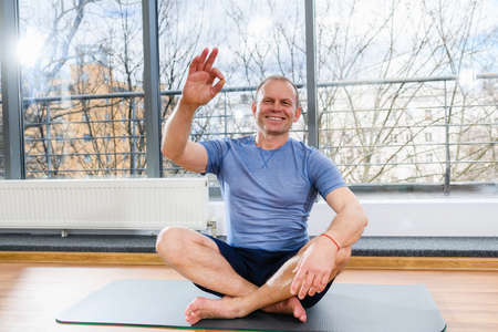 Positive sportive middle age man sit on floor mat and show okay gesture in light studio, healthy lifestyleの写真素材