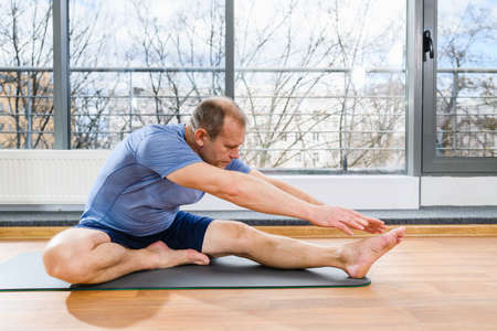 Middle age fit man sit on floor mat in light studio and stretch hands and feet, yoga fitness workout exercisesの写真素材