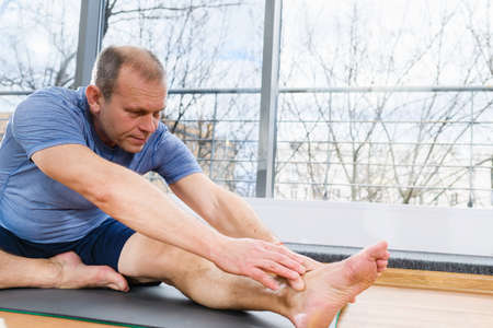 Middle age fit man sit on floor mat in light studio and stretch hands and feet, yoga fitness workout exercisesの写真素材