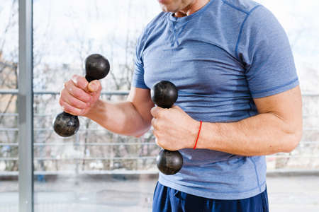 Sportive man torso in blue tight t-shirt with small dumbbells and red thread at light studio window backgroundの写真素材