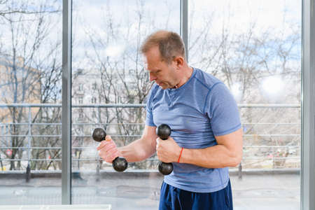 Middle age sportive fit smiling man in blue shirt workout with dumbbells in light gym with panoramic windowの写真素材