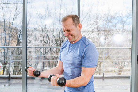 Middle age sportive fit smiling man in blue shirt workout with dumbbells in light gym with panoramic windowの写真素材