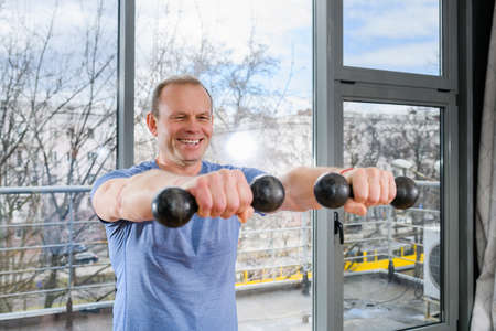 Smiling middle age fit sportsman exercise at light gym with panoramic window with weights, hands muscles training workoutの写真素材