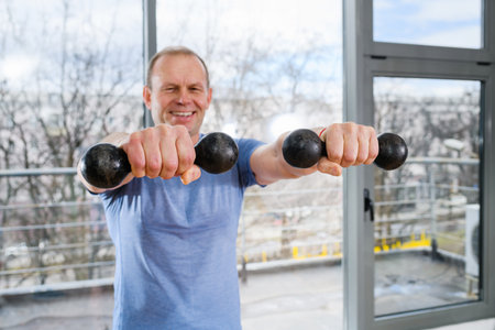 Smiling middle age fit sportsman exercise at light gym with panoramic window with weights, hands muscles training workout. Focus on hands with dumbbells.の写真素材