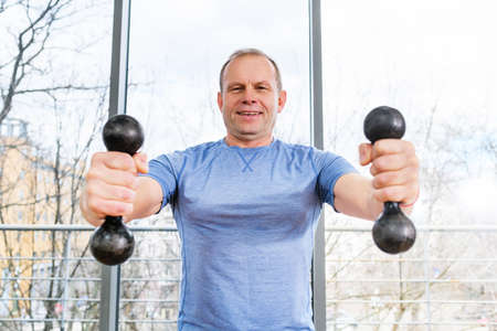 Smiling middle age fit sportsman exercise at light gym with panoramic window with weights, hands muscles training workoutの写真素材
