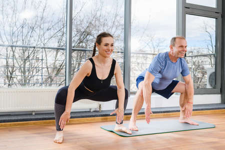 Young woman and mature man practice yoga poses in light studio with panoramic window, stay on mat with wide legs and hands downの写真素材
