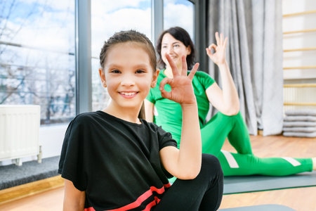 Sportive woman show okay gesture during fitness yoga exercises on mats near window, healthy family lifestyle. Focus on the girl.の写真素材